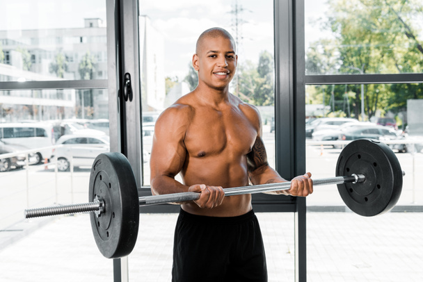 handsome muscular young sportsman holding barbell and smiling at camera in gym - Foto, Imagem