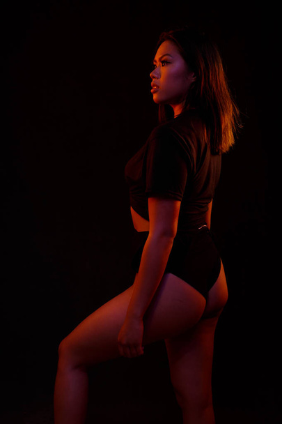 Fit young asian woman in black one piece poses against black under orange and blue colored lights - Фото, зображення