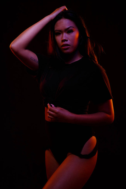 fit junge asiatische Frau in schwarz Einteiler posiert gegen Schwarz unter orangefarbenen und blauen Lichtern - Foto, Bild