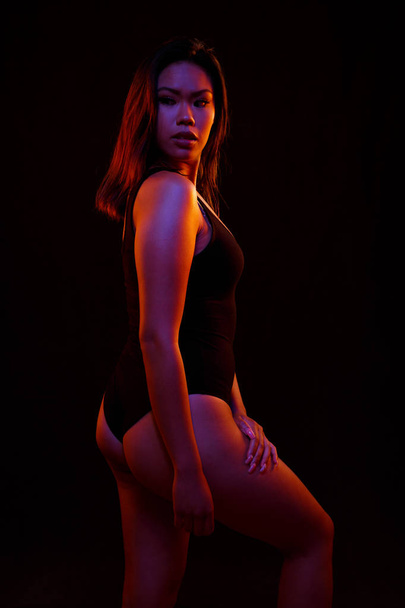 Fit young asian woman in black one piece poses against black under orange and blue colored lights - 写真・画像