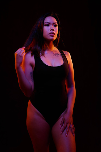 S'adapter jeune femme asiatique en noir pose une seule pièce contre noir sous les lumières de couleur orange et bleue
 - Photo, image