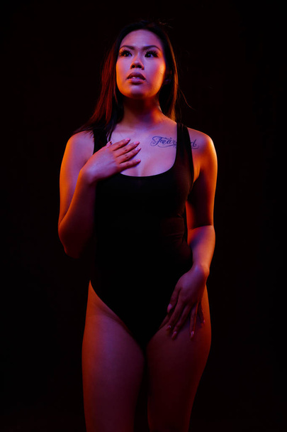 Fit young asian woman in black one piece poses against black under orange and blue colored lights - Photo, image