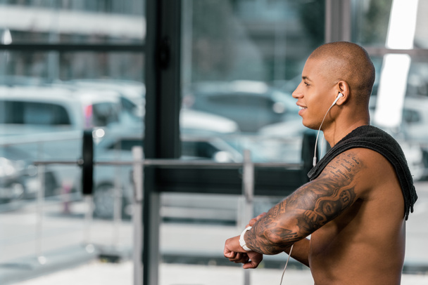 boční pohled na usmívající se shirtless sportovec v sluchátka pomocí hodinek smartwatch v tělocvičně  - Fotografie, Obrázek