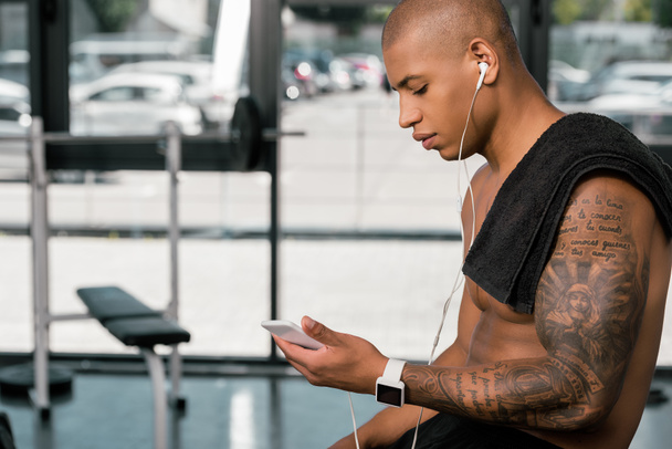 side view of muscular shirtless young man in earphones sitting and using smartphone in gym - Photo, Image