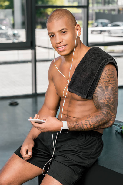 muscular young sportsman in earphones using smartphone and smiling at camera in gym - Φωτογραφία, εικόνα