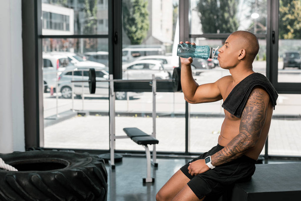 side view of muscular shirtless african american sportsman drinking water in gym - Photo, Image