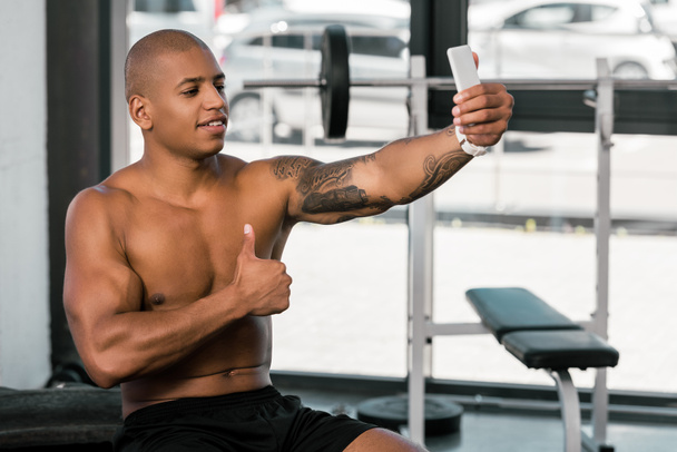 sonriente musculoso afroamericano hombre mostrando el pulgar hacia arriba y tomando selfie con smartphone en gimnasio
 - Foto, Imagen