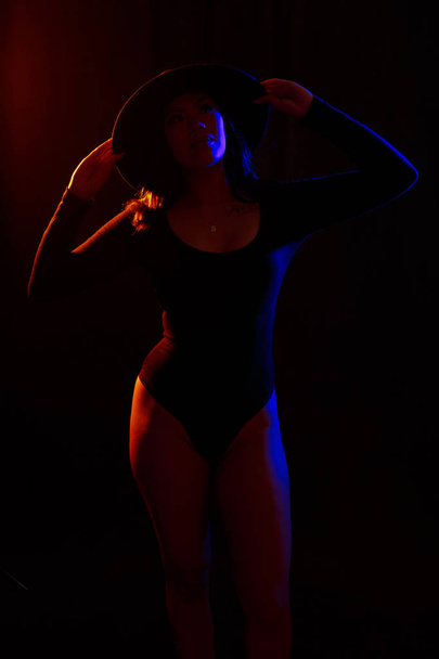 Fit jonge Aziatische vrouw in zwarte ééndelig en ketting vormt tegen zwart onder de oranje / blauwe gekleurde lampen - Foto, afbeelding