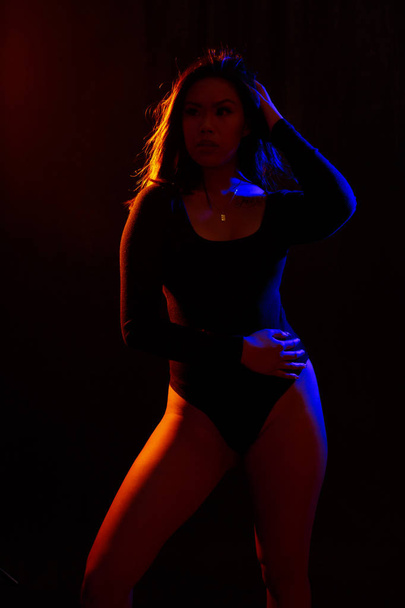 Fit young asian woman in black one piece and necklace poses against black under orange and blue colored lights - Fotó, kép