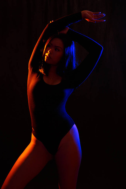 Fit young asian woman in black one piece and necklace poses against black under orange and blue colored lights - Foto, afbeelding