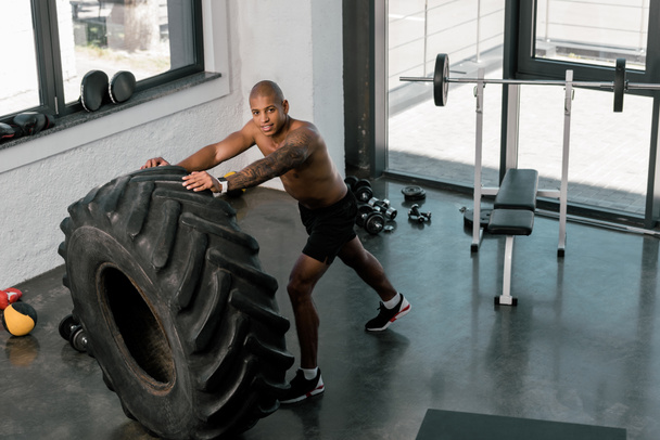 Blick aus der Vogelperspektive auf einen muskulösen jungen Mann, der im Fitnessstudio mit Reifen trainiert und in die Kamera lächelt - Foto, Bild