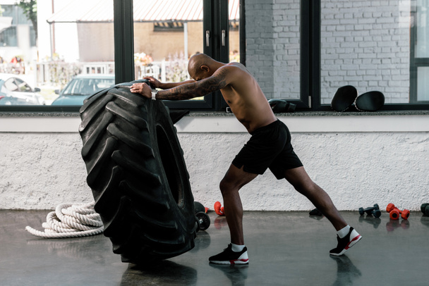 side view of muscular shirtless african american sportsman training with tire in gym - 写真・画像