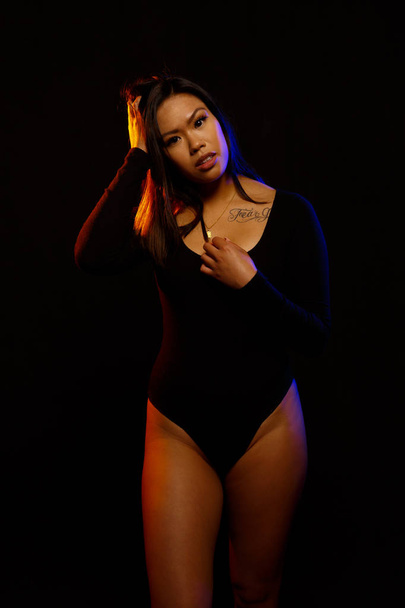 Fit young asian woman in black one piece and necklace poses against black under orange and blue colored lights - Zdjęcie, obraz