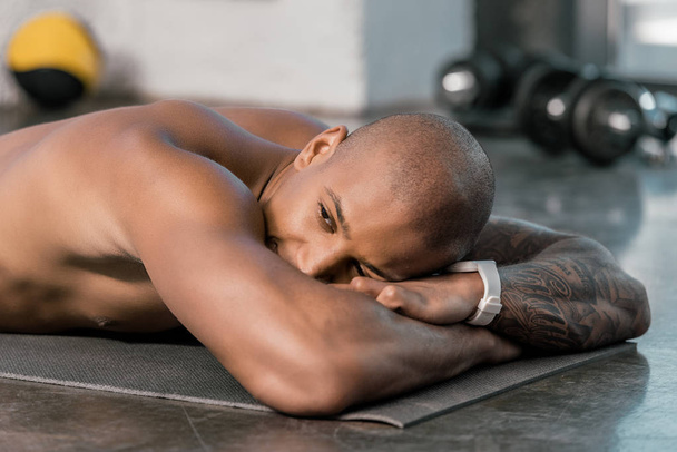 fröhlicher junger afrikanisch-amerikanischer Athlet ruht sich auf Fitnessmatte im Fitnessstudio aus - Foto, Bild