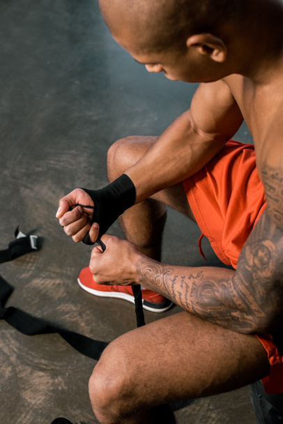 high angle view of tattooed african american sportsman wrapping hand in boxing bandage at gym - Фото, зображення