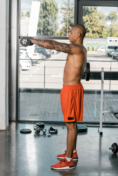boční pohled na svalové shirtless africké americké sportovce cvičení s činkami v posilovně - Fotografie, Obrázek