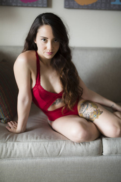 Young caucasian woman sits on couch in natural light while wearing red lingerie - Photo, Image