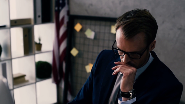 όμορφος επιχειρηματία σε κοστούμι και γυαλιά, κρατώντας το χέρι στο πηγούνι και σκέψης ενώ κάθεται στο χώρο εργασίας - Πλάνα, βίντεο