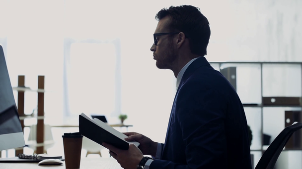 vista lateral del hombre de negocios guapo en ropa formal y gafas con cuaderno y trabajando en la oficina
 - Metraje, vídeo