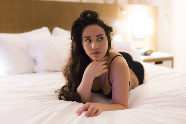 Young caucasian woman wearing see through black lingerie relaxes on a bed with white sheets under studio lighting - Fotó, kép