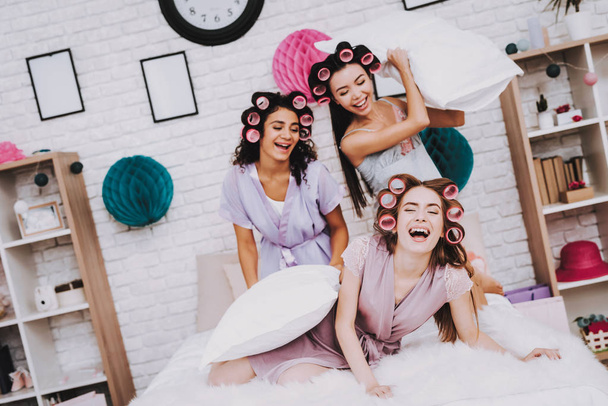 Fight Pillows. Home Party. Women in Bathrobes. White Pillows. Bright Room. White Interior. Beautiful International Woman. Smiling Girls. White Pillow in Hand. Funny People. Women with Pink Curlers. - Photo, image
