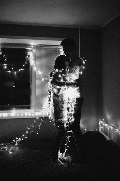 Loving couple hug each other  in garland in bedroom. Black and white photo with noise filter - Фото, изображение