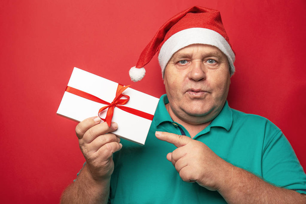 Portrait of man in red Christmas hat pointing with finger on Gift Box on red background, New Year presents for family - Photo, Image