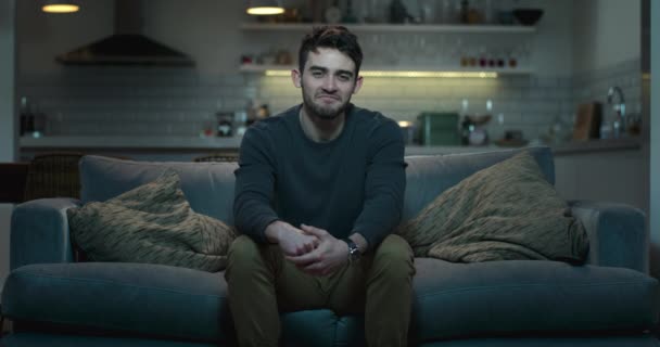 junger Mann sitzt auf einer Couch in einem abgedunkelten Wohnzimmer, lächelt und lacht über etwas im Fernseher. - Filmmaterial, Video