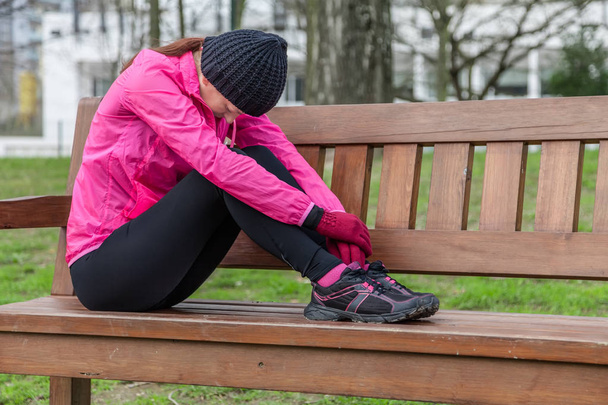 Nuori urheilija nainen väsynyt tai masentunut lepää penkillä kylmänä talvipäivänä kaupunkipuiston radalla.
. - Valokuva, kuva
