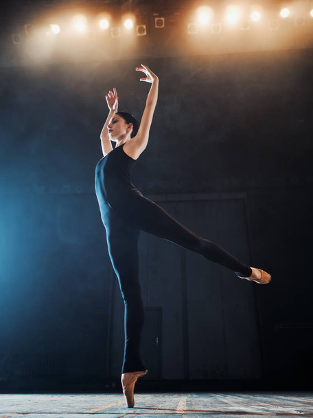 Young beautiful ballerina on smoke stage dancing modern ballet. performs smooth movements with hands against spotlights background. Woman in black costume on scene - Fotó, kép