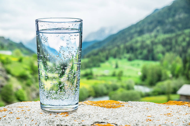 Glass of water on the stone. Blurred snow mountains tops and green forests at the background, as a symbol of freshness and purity. - Photo, Image