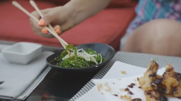 Nainen maistaa japanilaista ruokaa. Lähikuva houkutteleva nuori valkoihoinen nainen puree merilevää salaattia ottaen sen syömäpuikoilla avoimella terassilla japanilainen ravintola
. - Materiaali, video