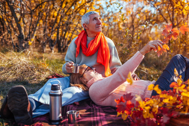 Senior couple having tea in autumn forest. Happy man and woman enjoying picnic and nature. Smiling man pouring tea out of thermos - Foto, afbeelding