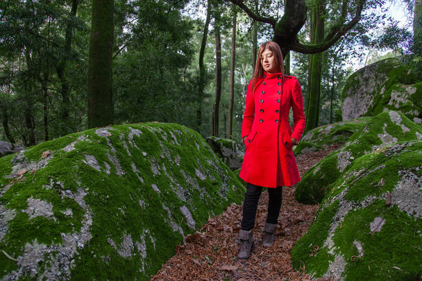 junge Frau, die traurig ist, wenn sie im Winter unter einem Sonnenstrahl in einem roten Mantel durch den Wald geht - Foto, Bild