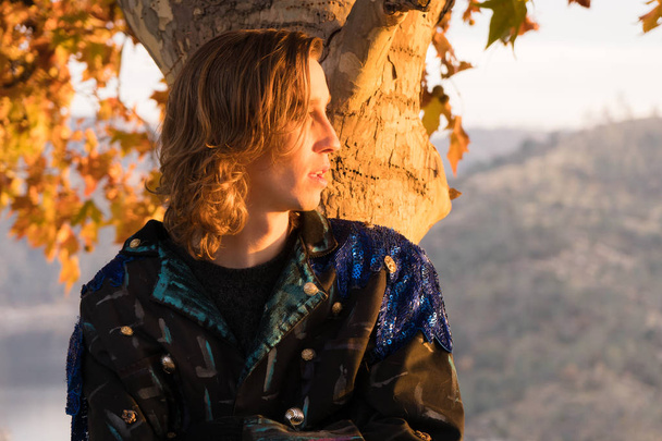 Young man with long hair poses against tree at sunset above scenic lake - Photo, Image