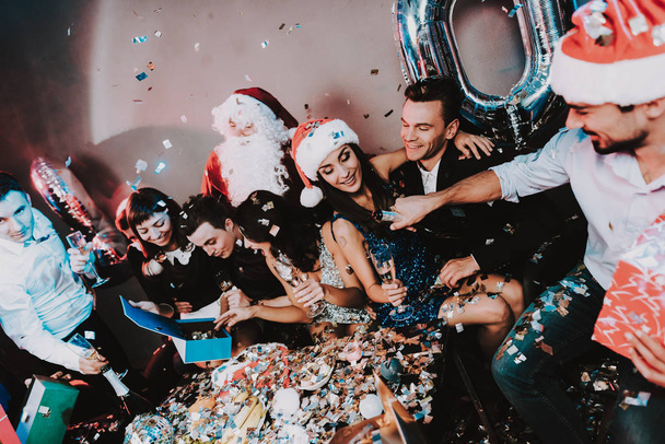 Young Happy People Celebrating New Year at Table. Santa Claus Costume. People in Red Caps. Happy New Year Concept. Bottle of Champagne. Celebrating of New Year. Man with Gift Box. - Fotoğraf, Görsel