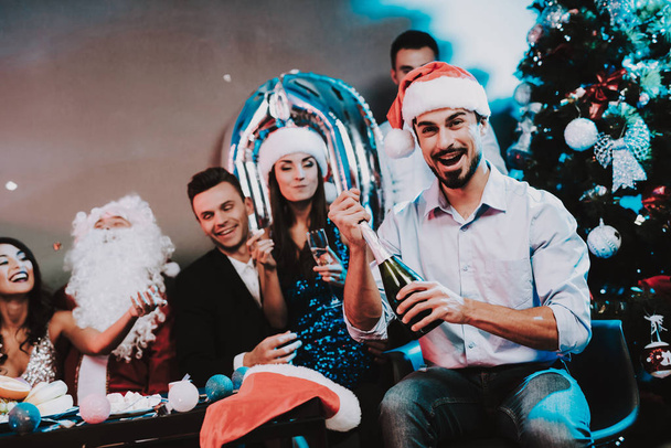 Bearded Man in Rad Cap Opening Bottle of Champagne. Santa Claus Costume. People in Red Caps. Happy New Year Concept. Bottle of Champagne. Celebrating of New Year. Young Woman in Dress. - Foto, Bild