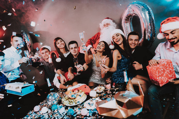 Young Happy People Celebrating New Year at Table. Santa Claus Costume. People in Red Caps. Happy New Year Concept. Bottle of Champagne. Celebrating of New Year. Man with Gift Box. - Foto, imagen