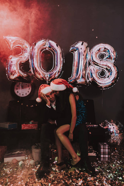Woman with Sad Man in Red Cap on New Year Party. Happy New Year. Indoor Party. Celebrating of New Year. Young Woman in Dress. Young Man in Suit. Upset Man. Santa Claus Cap. Sitting Man. - Foto, Bild