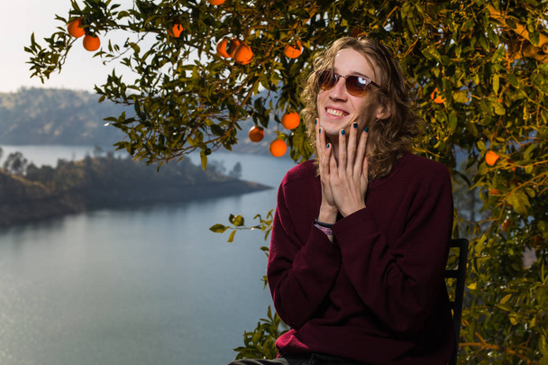 Young caucasian man with long hair and casual clothing poses under an orange tree with lake in background - Photo, Image