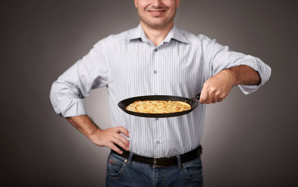 man poseren met een pannenkoek in een pan, wit overhemd en broek, grijze achtergrond, ondiepe scherptediepte, scherpe pannenkoek en wazig gezicht - Foto, afbeelding