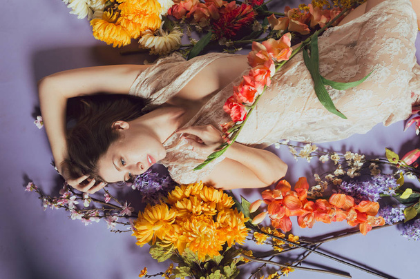 Young caucasian woman lies in a pile of flowers against a blue background. Sheer white dress - Φωτογραφία, εικόνα