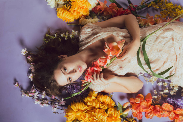 Молода біла жінка лежить в купі квітів на синьому фоні. Біла сукня
 - Фото, зображення