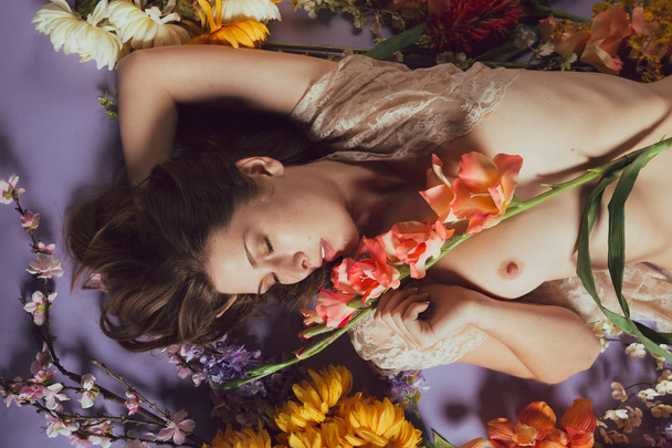 Young caucasian woman lies in a pile of flowers against a blue background. Sheer white dress - 写真・画像