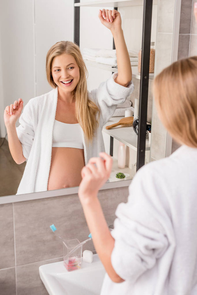 happy attractive pregnant woman in bathrobe dancing in front of mirror in bathroom - Фото, изображение