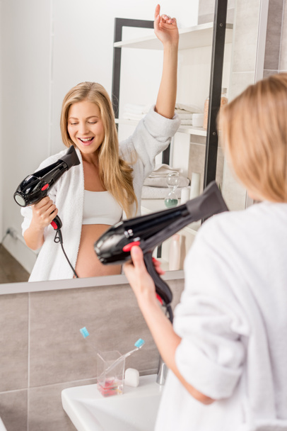 attractive happy pregnant woman in bathrobe singing at hair dryer in front of mirror in bathroom - Foto, afbeelding