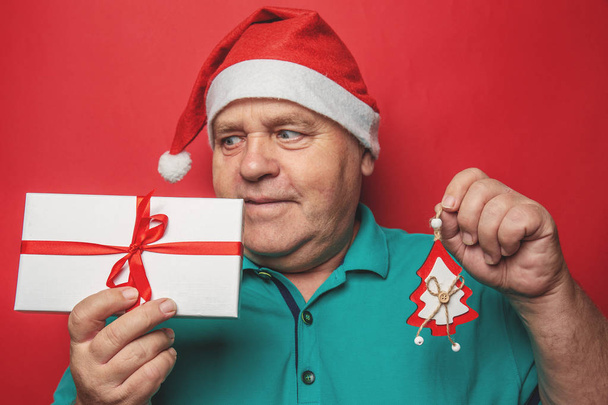 Funny man in red Christmas hat holds in hands gift box and New Year tree toy, presents for relative people and family on holidays - Valokuva, kuva