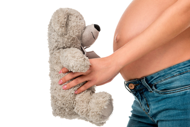 partial view of pregnant girl holding teddy bear in front of belly isolated on white - Fotoğraf, Görsel