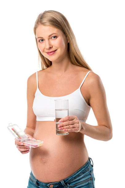 usmívající se těhotná žena držící krabička s vitamíny a sklenicí vody izolované na bílém - Fotografie, Obrázek