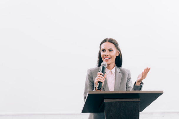 aantrekkelijke lachende docent in de microfoon praten en gebaren op podium tribune tijdens seminar in de conferentiezaal - Foto, afbeelding